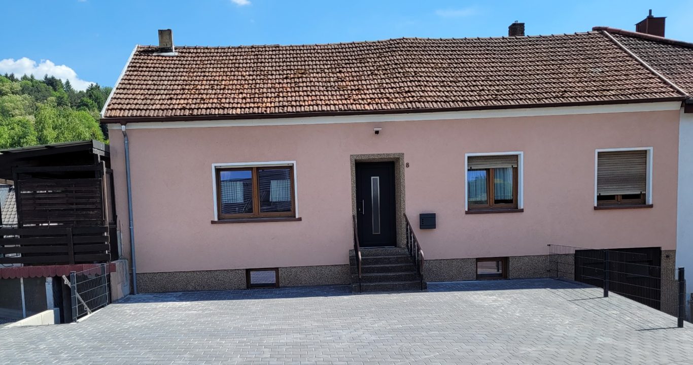 Einfamilienhaus mit rosa Fassade und schwarzer Eingangstür, umgeben von grünen Hügeln.